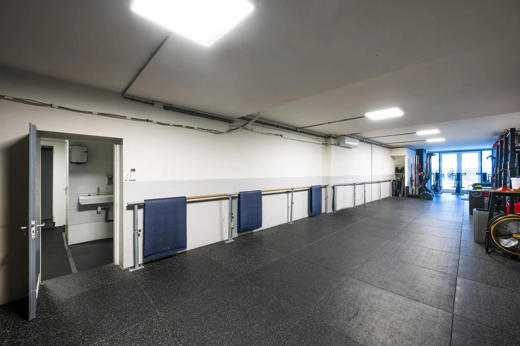 Fitnessruimte aan de Albert Cuypstraat met rubberen vloer, wandrekken, fitnessapparatuur en een open deur naar een toiletruimte.